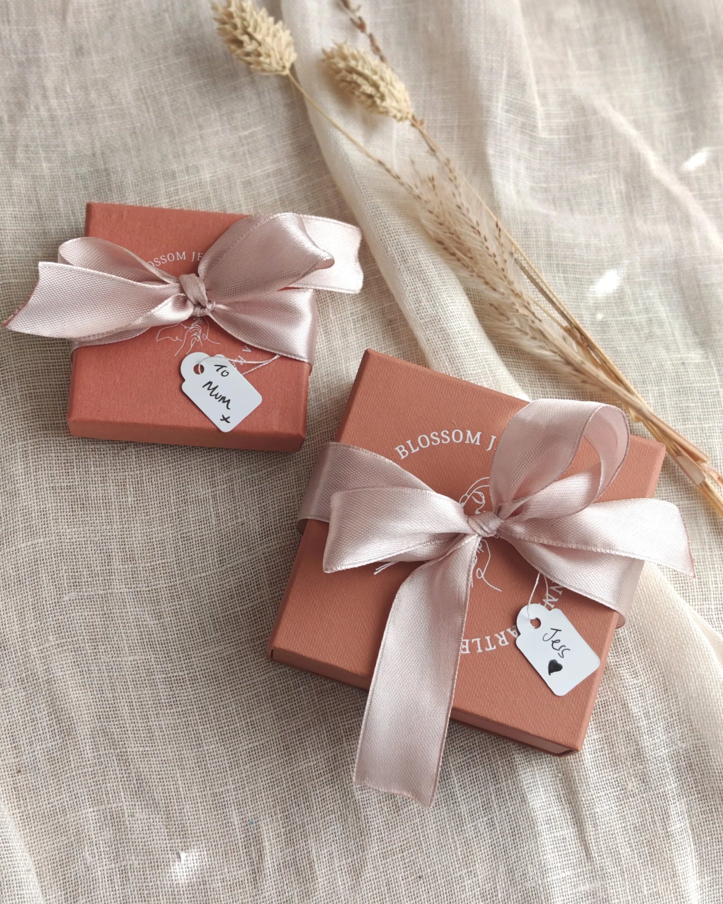 Two pink jewelry gift boxes with satin bows and custom tags on linen, dried flowers nearby