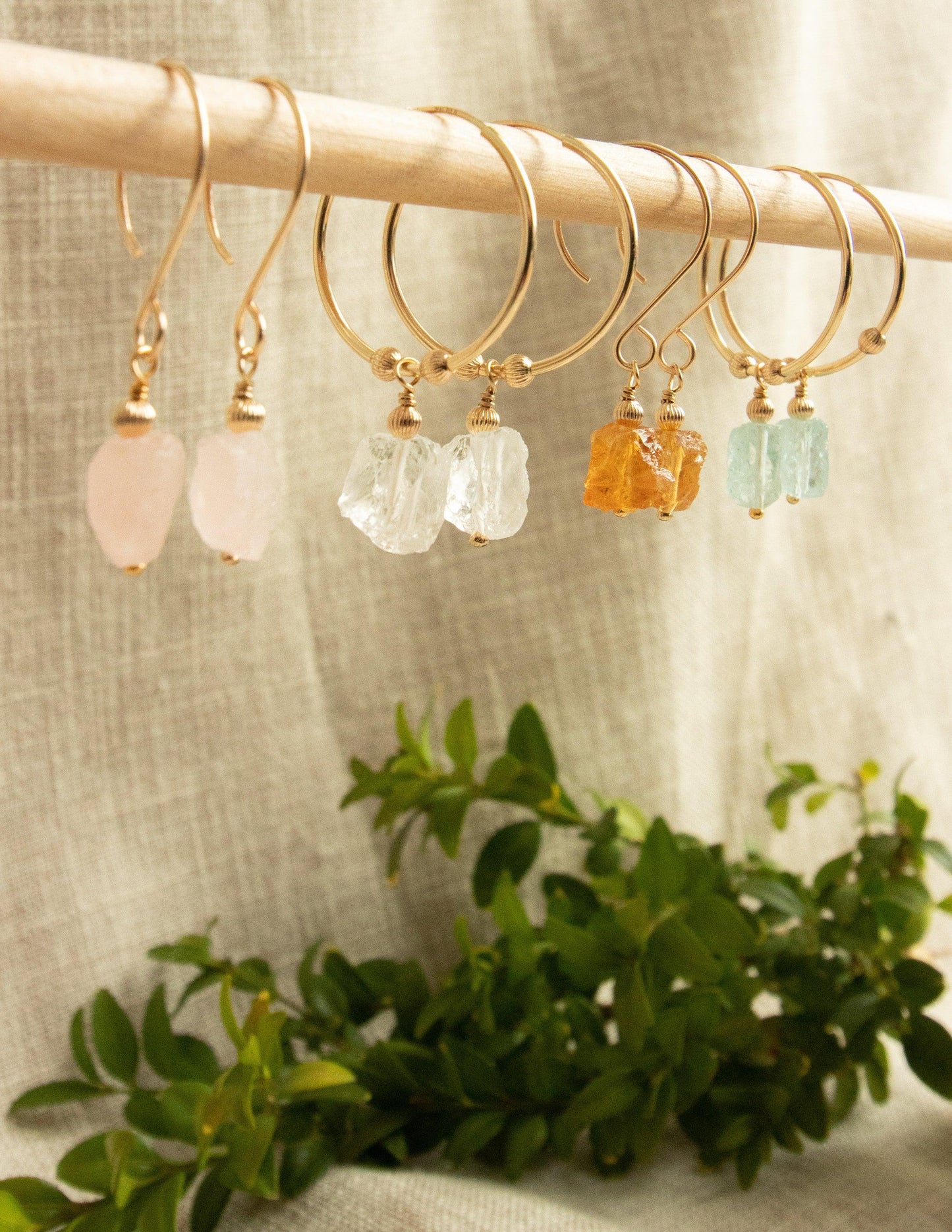 Gold hoop earrings with raw gemstone drops in pastel colors, displayed on a wooden rod
