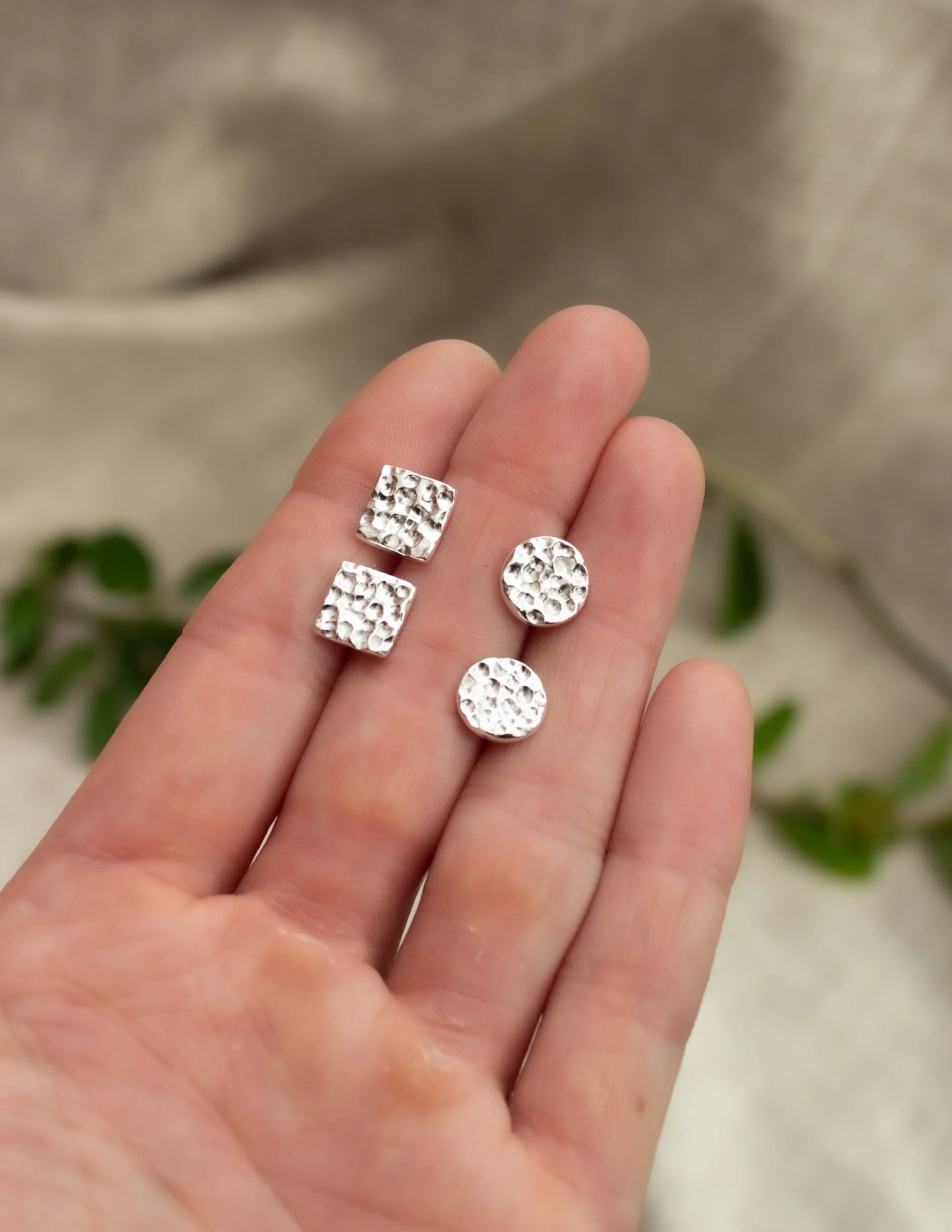 Hand holding hammered sterling silver stud earrings, round and square shapes, on neutral background.