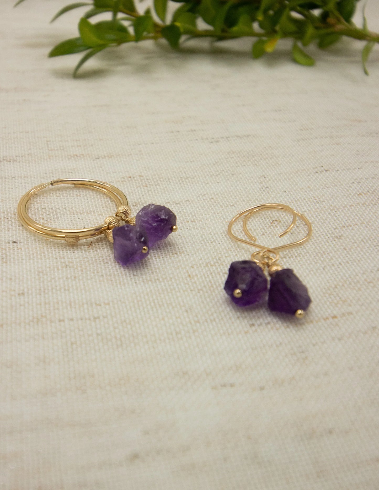 Gold hoop earrings with raw amethyst gemstone dangles on linen, green plant background