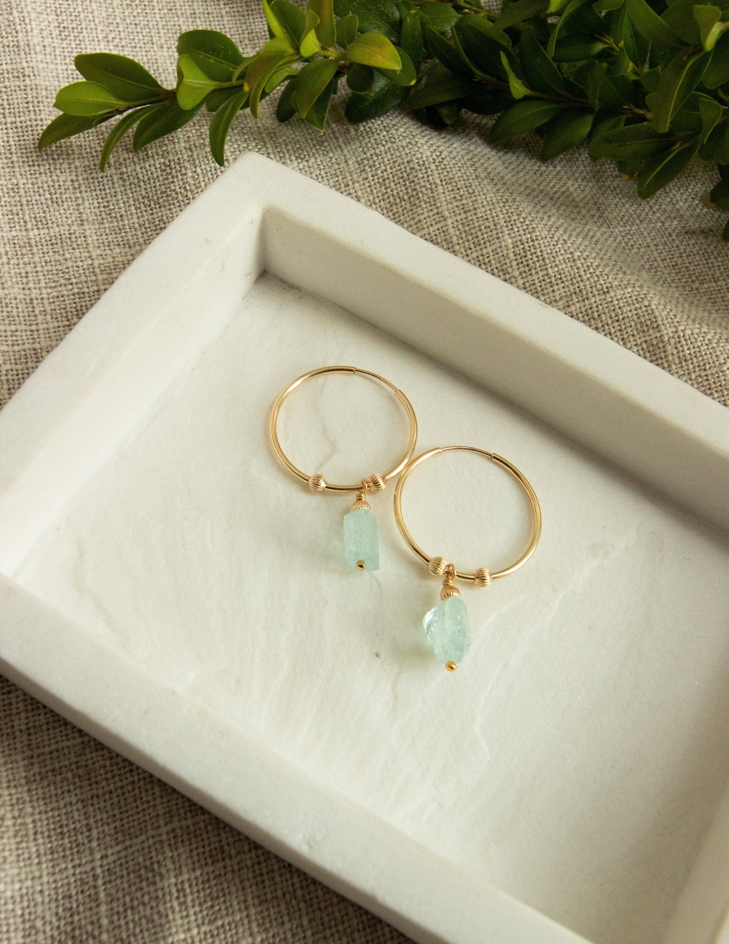 Gold hoop earrings with raw aquamarine gemstone drops on a ceramic tray