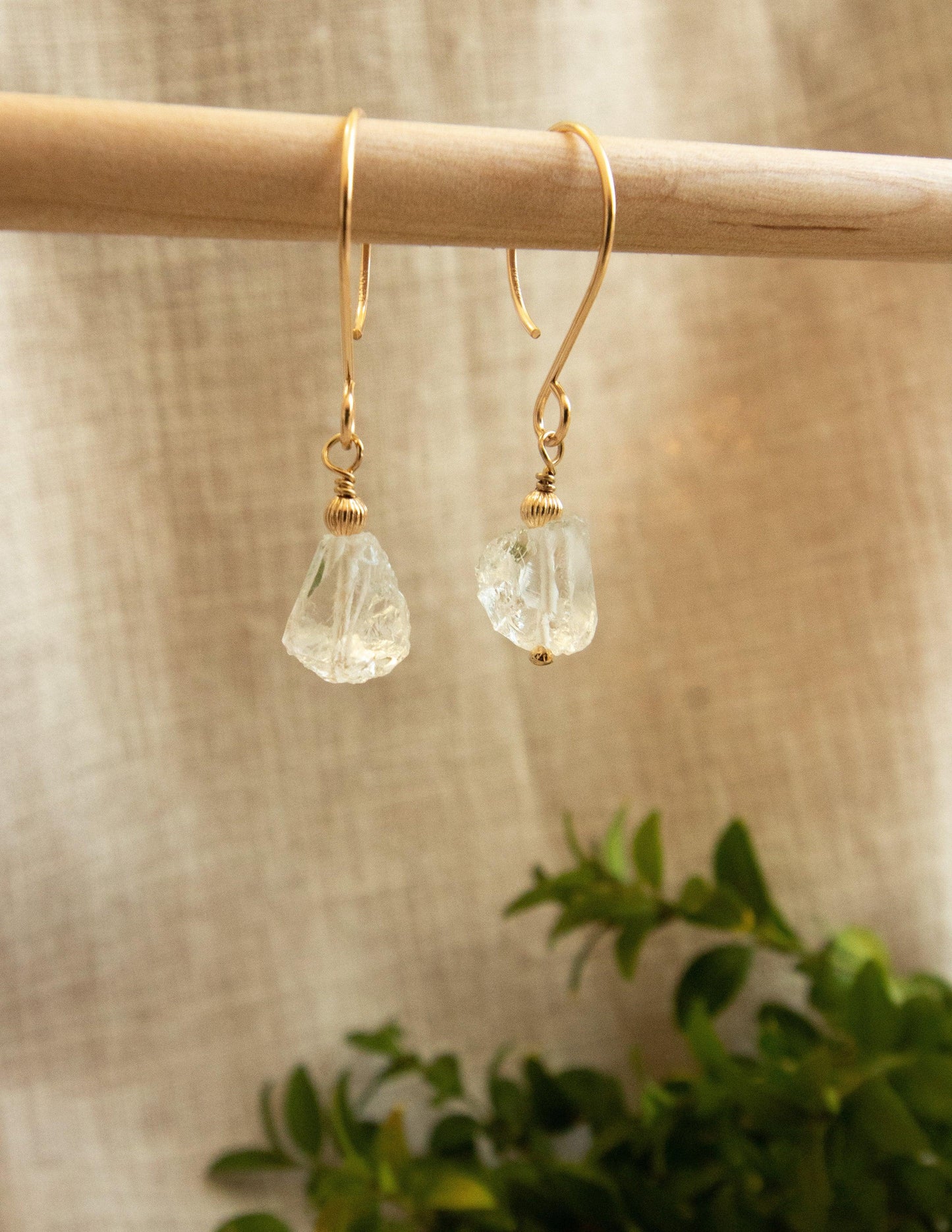 Gold filled earrings with raw clear gemstones hanging on a wooden rod, beige linen background
