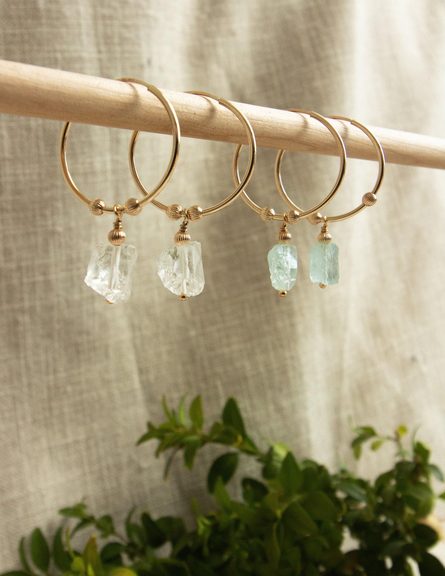 Gold hoop earrings with raw gemstone dangles, displayed on a wooden rod with a neutral background.