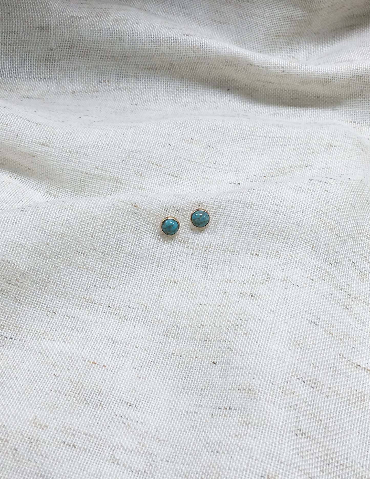 Turquoise gemstone stud earrings with gold setting on white fabric background